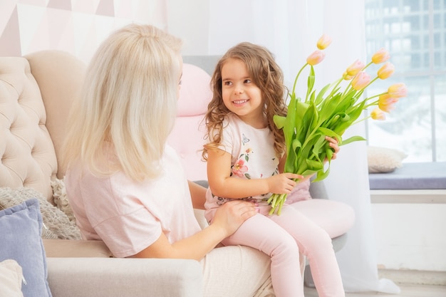 Happy mothers day. Child daughter congratulates mom and give her bouquet of flowers tulips in pink livingroom in their house.