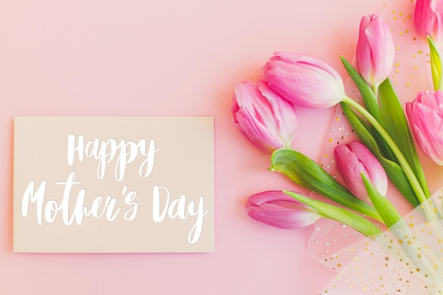 Happy mothers day text on greeting card and pink tulips on pink\
background stylish greeting card happy mother\'s day gratitude and\
love to mom handwritten lettering