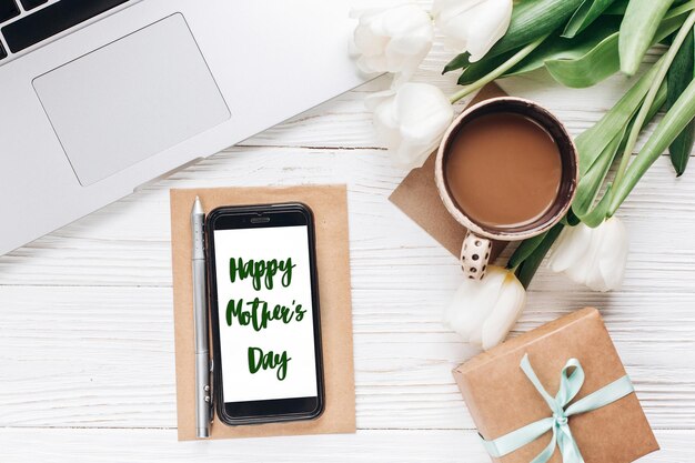 Happy mothers day text sign on phone screen and laptop with morning coffee and tulips on white wooden rustic background stylish flat lay with flowers and working gadgets with space for text