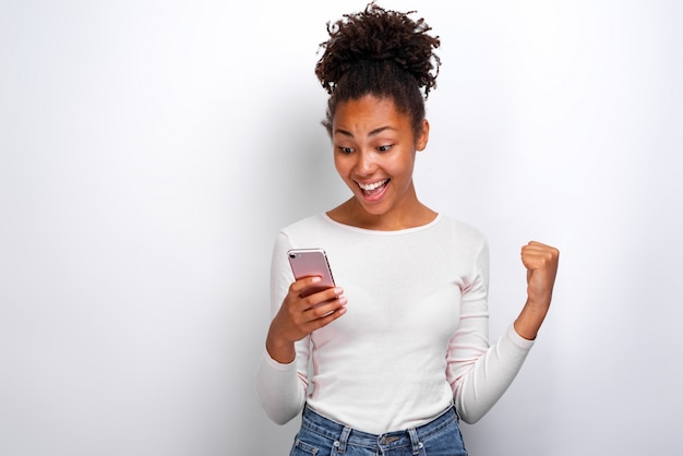 Happy mulatto woman holding a smartphone in her hand and screaming with winner gesture looking at the screen