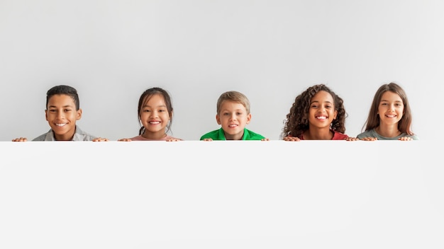 Happy multicultural children posing with empty white poster gray background