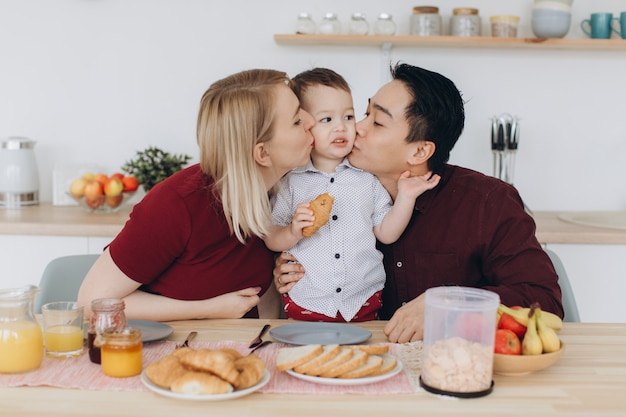 Happy multicultural family. Asian dad and his caucasian blonde wife have breakfast with their beautiful son in the kitchen.