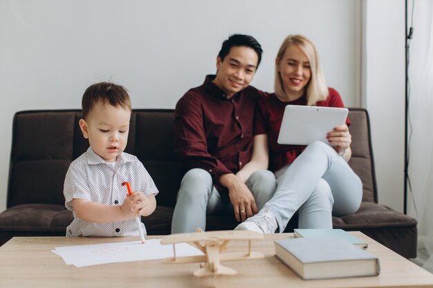 Happy multicultural family. Asian man and Caucasian woman with their son watching video on tablet and phone.