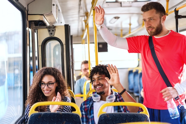 Happy multicultural friends waving to someone while sitting and standing in city bus.