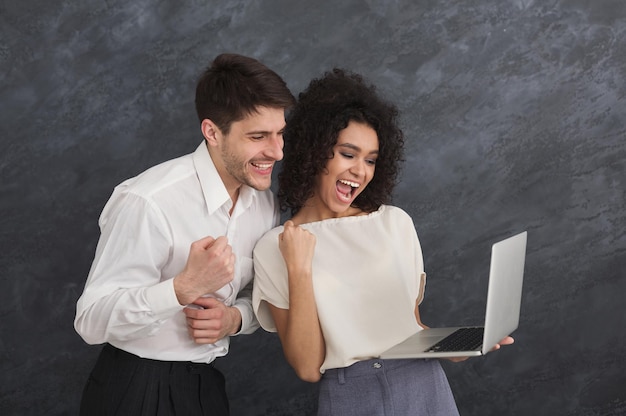 Happy multiethnic couple in formal clothes using laptop, celebrating success on grey background, copy space