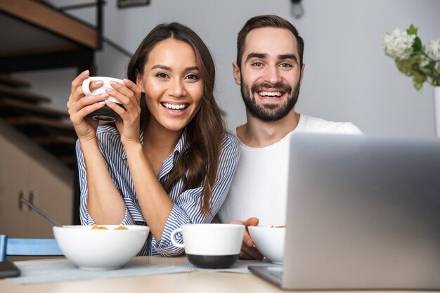 ラップトップコンピューターを見て、キッチンで朝食をとっている幸せな多民族のカップル