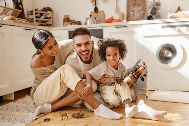 Happy multiethnic family parents and son playing with toy plane\
on floor in cozy kitchen at home