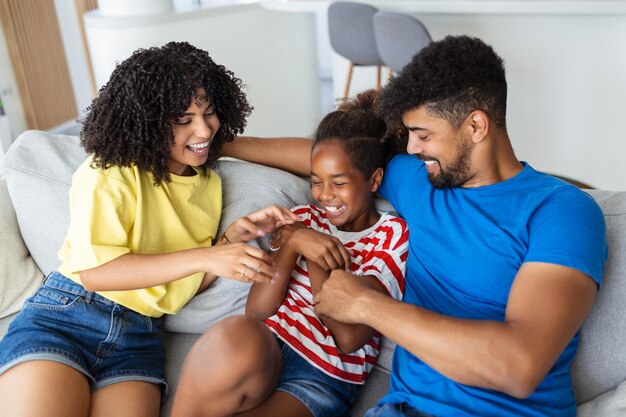 Photo happy multiethnic family sitting on sofa laughing together cheerful parents playing with their daughter at home father tickles his little girl while the mother is smiling