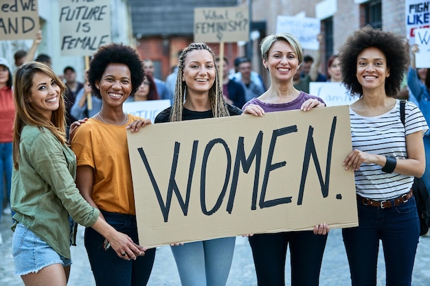 Happy multiethnic group of females holding banner with 'women' inscription while protesting for their rights on city streets