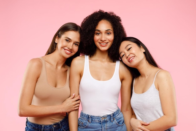 Happy multiethnic young women posing and smiling to camera standing over pink background studio shot