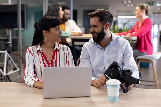 Happy multiracial business colleagues looking at each other while sitting with vr headset and laptop