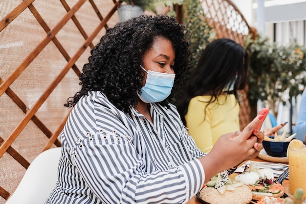 Happy multiracial friends using mobile phones outdoors at brunch restaurant while wearing masks - Focus on african american girl