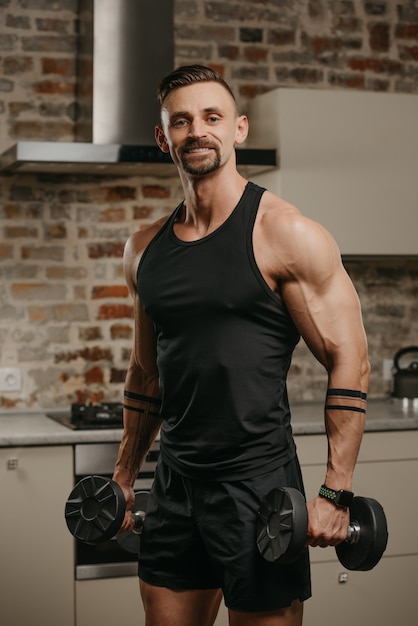 A happy muscular man with a beard is posing with dumbbells in his apartment