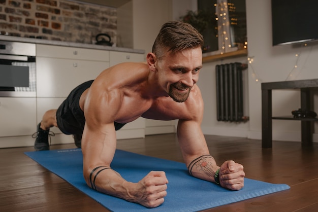 A happy muscular man with a naked torso is doing a plank on a blue yoga mat in his apartment in the evening