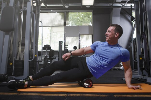 Happy muscular sportsman stretching muscles using foam roller at sports studio