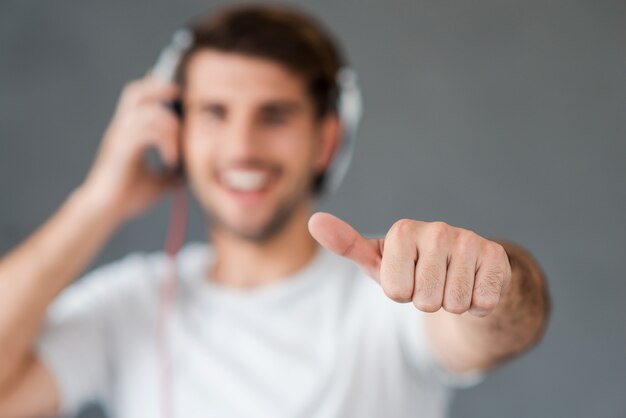 Happy music lover. Happy young man in headphones showing his thumb while standing against grey background