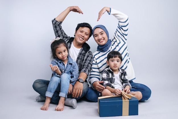 회색 배경에 고립 된 팔로 심장 기호를 만드는 바닥에 앉아있는 행복한 무슬림 가족