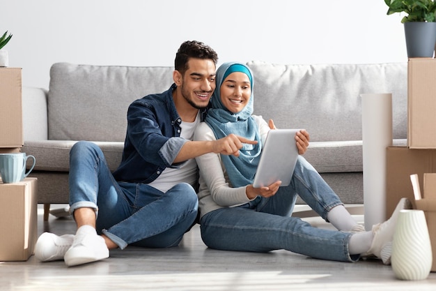 Happy muslim family sitting on floor in new apartment with digital tablet among boxes with belongings, buying furniture for house or working on design together, drinking coffee and having conversation