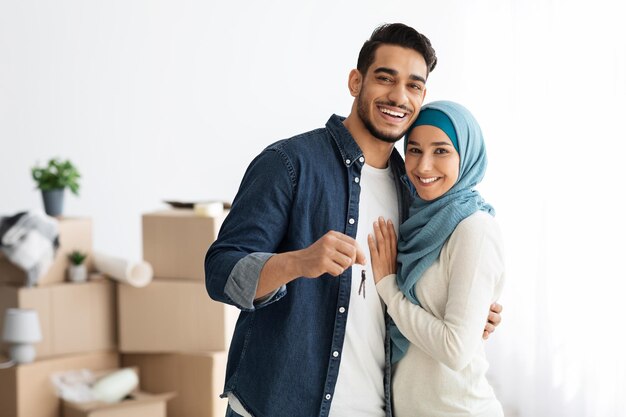 Happy muslim family standing over paper boxes and showing keys from their new apartment. Handsome arab man and young woman in hijab moving to their house, unpacking belongings, copy space