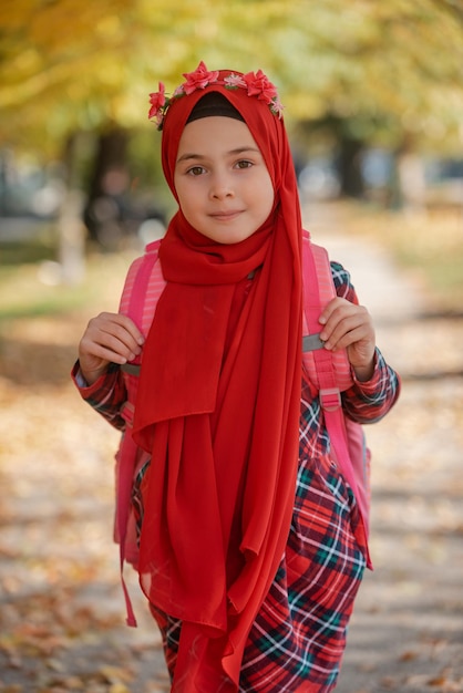 秋の公園を歩く学校のバックパックとファッション ヒジャブで幸せなイスラム教徒の少女。