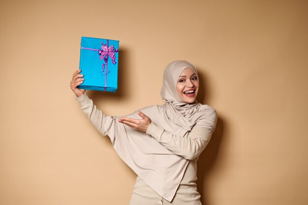 선물로 그녀의 손을 들고 카메라에 제시하는 hijab의 행복 무슬림 여성.