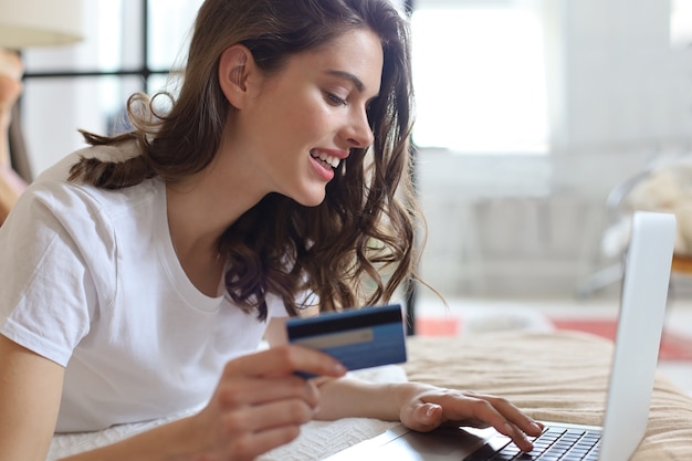 Happy natural brunette using credit card and laptop in bedroom.