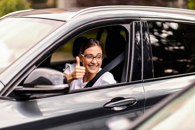 A happy nerdy girl is driving a car and showing thumbs up to other drivers