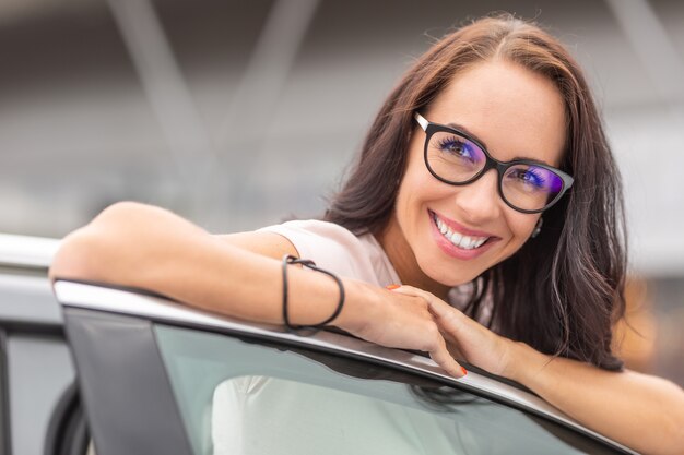 행복한 새 여성 자동차 소유자는 큰 미소로 차량의 열린 문에 기대어 있습니다.