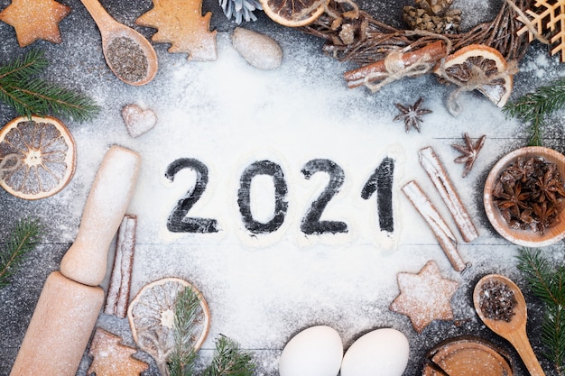 Happy New Year 2021 written on flour. Christmas tree branches, gingerbread cookies, spices and baking supplies on black wood surface