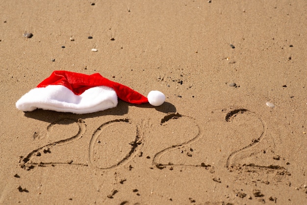 Felice anno nuovo 2022, scritte sulla spiaggia. numeri 2022 anno in riva al mare con un berretto di capodanno, messaggio scritto a mano. foto di alta qualità