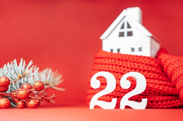 Felice anno nuovo 2022. i numeri in legno bianco 22 stanno con una casa giocattolo bianca avvolta in una sciarpa rossa e un ramo di un albero di natale su sfondo rosso. buon natale. copia spazio