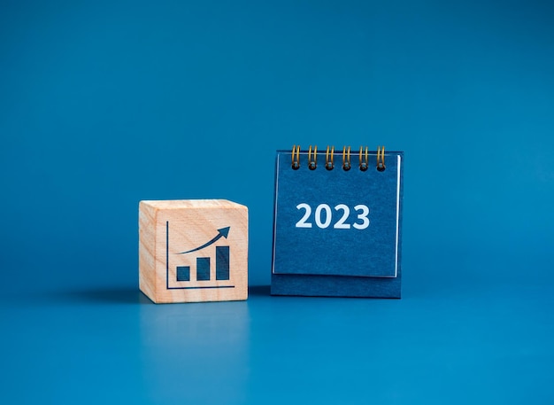 새해 복 많이 받으세요 2023 배경 2023 숫자 연도는 파란색 배경 미니멀리스트에 격리된 성장하는 그래프 아이콘이 있는 나무 큐브 블록의 작은 책상 달력에 새 목표를 시작할 준비가 되었습니다.