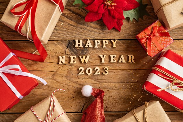 Happy new year 2023 festive background with wooden letters and craft gift boxes, poinsettia and santa hat on wooden background.