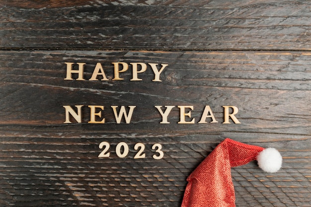 新年あけましておめでとうございます 2023年グリーティング カード赤いサンタ帽子お祭りの背景を持つ木製の背景に木製の文字で作られた引用
