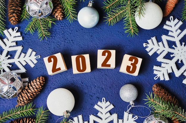 Happy new year 2023 text sign on wooden cubes with frame of christmas tree branch and cones on blue background Snowflakes