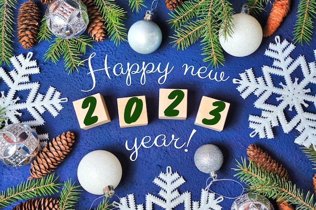 Happy new year 2023 text sign on wooden cubes with frame of\
christmas tree branch and cones on red background snowflakes