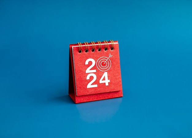 Happy new year 2024 banner background 2024 numbers year with target dart icon on red small desk calendar cover standing on blue background minimal style Business goals plan and success concepts