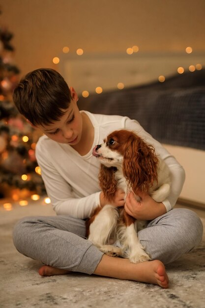 写真 新年あけましておめでとうございます 軽い自家製のパジャマを着た少年が クリスマスツリーの近くの寝室で ペットのカヴァリアー・キング・チャールズ・スパニエルを抱きしめています