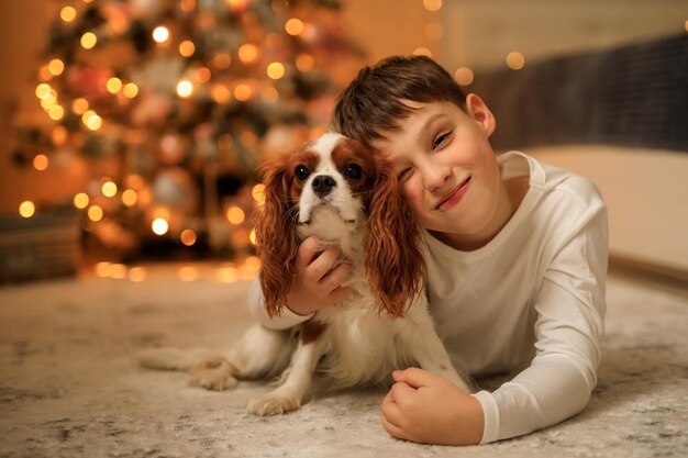 Happy New Year a boy in light homemade pajamas hugs his pet cavalier king charles spaniel at home in the bedroom near the Christmas tree