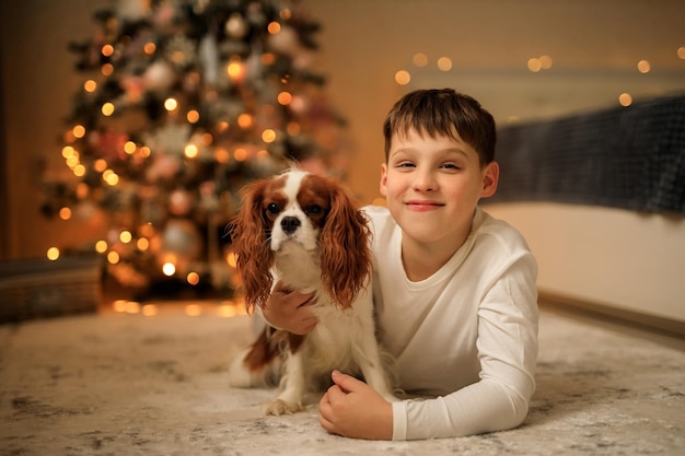 Foto buon anno un ragazzo in leggero pigiama fatto in casa abbraccia il suo animale domestico cavalier re charles spaniel a casa nella camera da letto vicino all'albero di natale