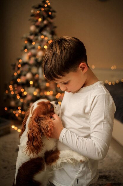 Foto buon anno un ragazzo in leggero pigiama fatto in casa abbraccia il suo animale domestico cavalier re charles spaniel a casa nella camera da letto vicino all'albero di natale