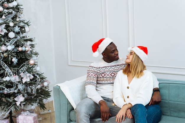 Happy New Year and Merry Xmas at home together. Smiling young african american guy and caucasian lady in Santa Claus hats, hug and look at camera in living room interior with tree and garlands.