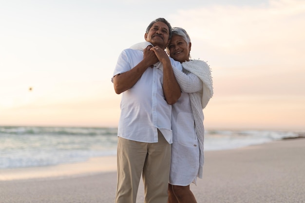 Happy newlywed senior multiracial couple embracing at beach against sky during sunset