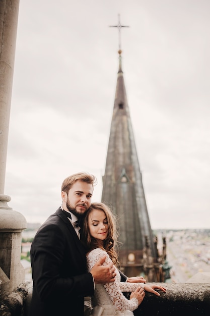 Photo happy newlyweds beautiful bride and stylish groom are hugging on the balcony of old gothic cathedral