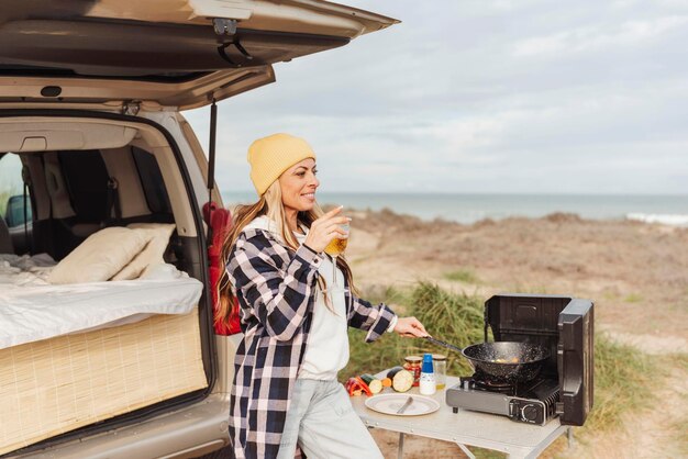 ビーチのコンセプト旅行と遊牧民のライフ スタイルの横にあるキャンピングカーの外で料理しながらビールのグラスを持つ幸せな遊牧民の女性