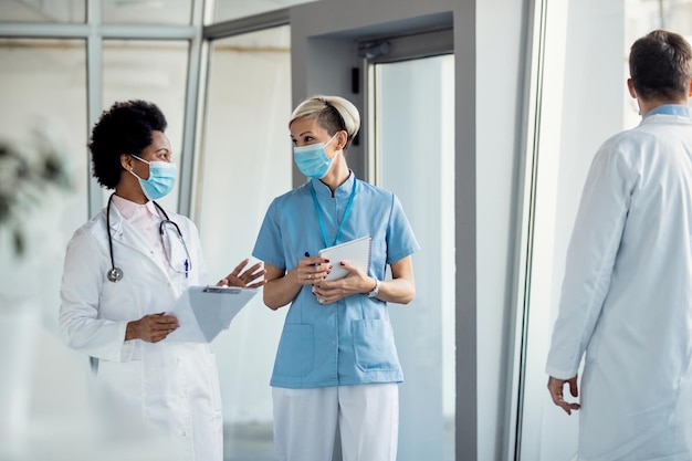 Happy nurse and black female doctor wearing face masks while\
communicating in a lobby at the hospital