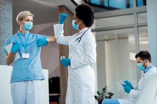 Happy nurse elbow bumping with female doctor while greeting in a lobby at medical clinic