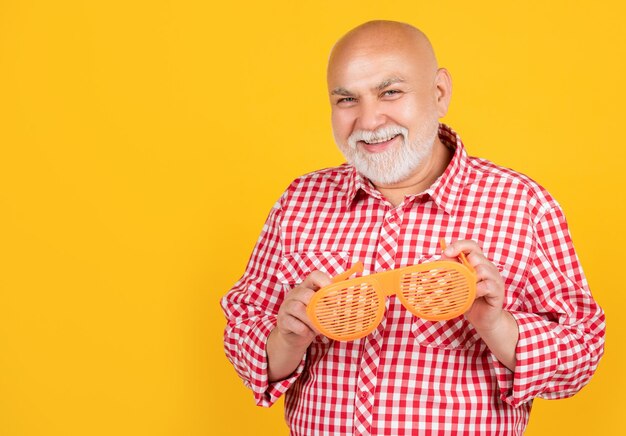 Happy old aged man in checkered shirt and party glasses on yellow background