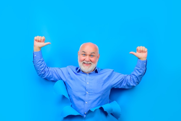 Happy old bearded man through hole in blue paper old man shows thumbs up copy space for advertising