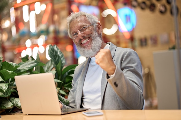 Счастливый старый бизнесмен празднует хорошие результаты, выигрывая деньги онлайн-ставкой